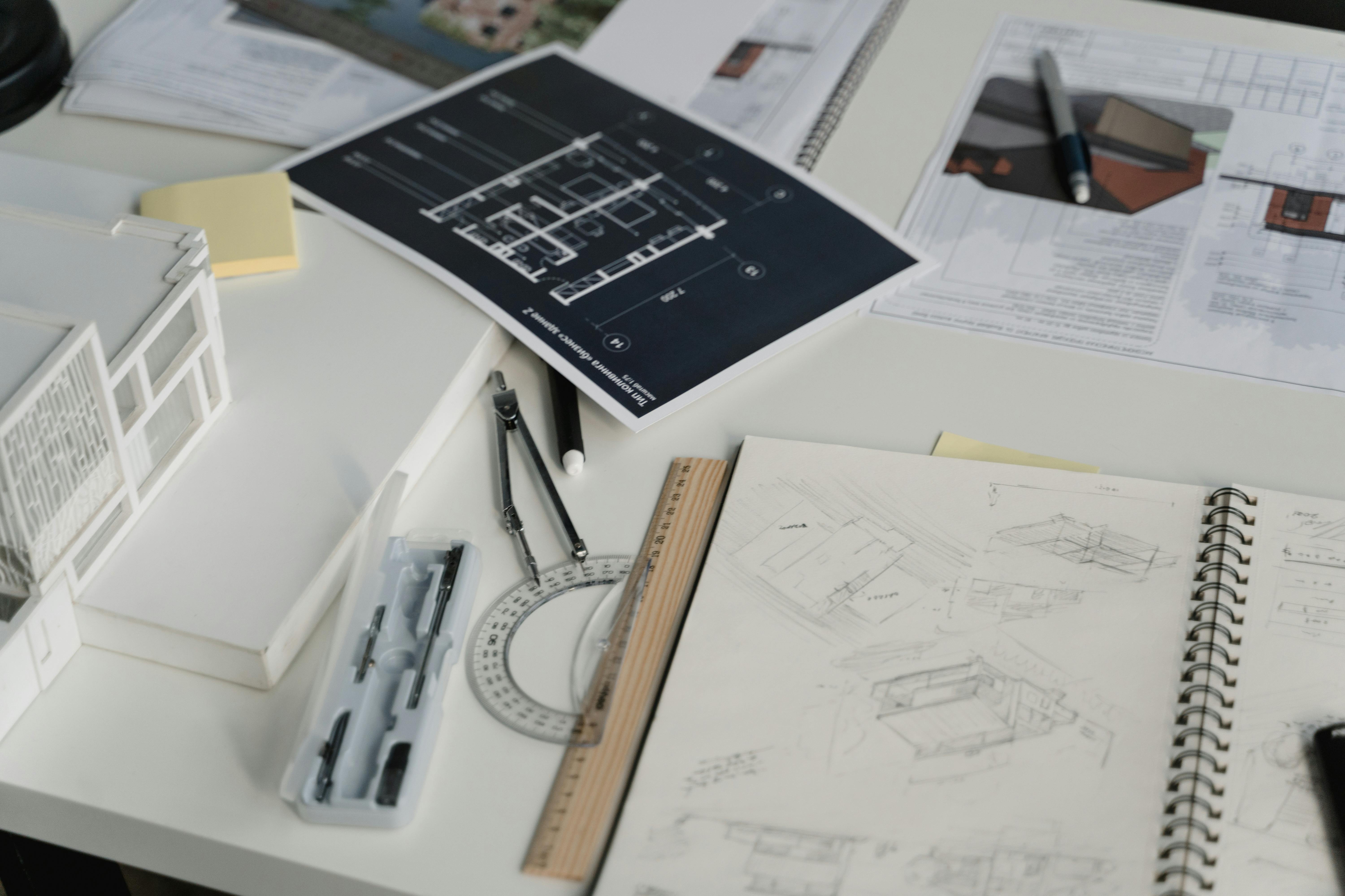 A workspace showing architectural plans and sketches, including a model building, drafting tools, pens, and notes on a table.
